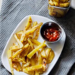 Crispy seasoned French fries served with ketchup on a white plate, garnished with a side of additional fries in a metal basket.