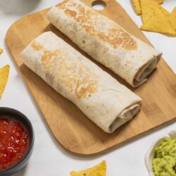 Freshly made chicken burritos on a wooden cutting board with chips and dips.