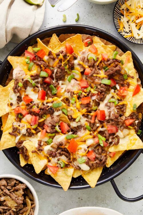 Crispy beef nachos with melted cheese, tomatoes, and green onions on black plate.
