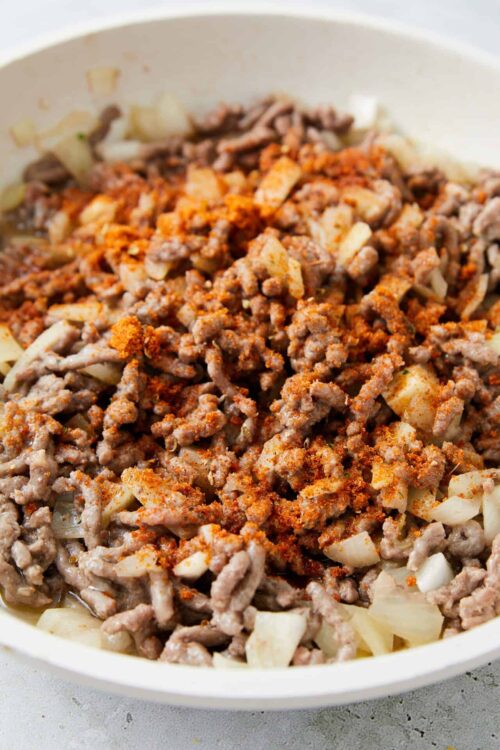 Ground beef and onion mixture in a white bowl, topped with spices and seasonings.