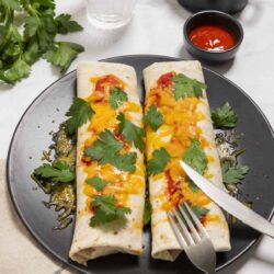 Crispy chicken burritos with melted cheese and fresh cilantro on a black plate.