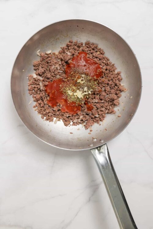 Ground beef being cooked with tomato sauce and seasonings in a stainless steel skillet.