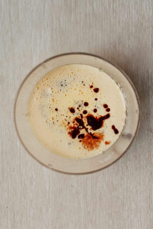 Creamy vanilla latte topped with chocolate syrup, in a clear glass on a neutral background.