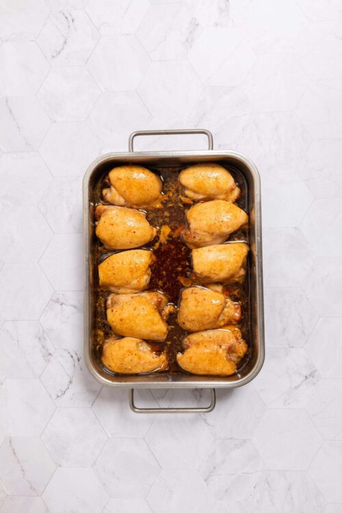 Juicy baked chicken thighs in a roasting pan on white marble background.