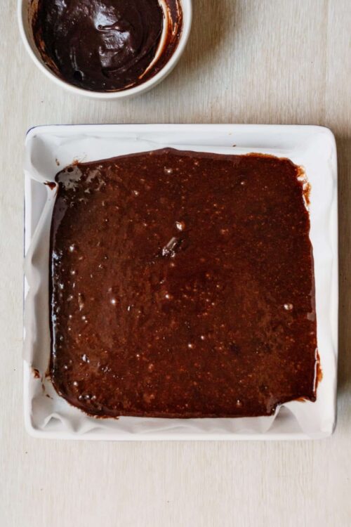 Decadent chocolate brownie batter in a white baking dish with chocolate ganache topping in a bowl.