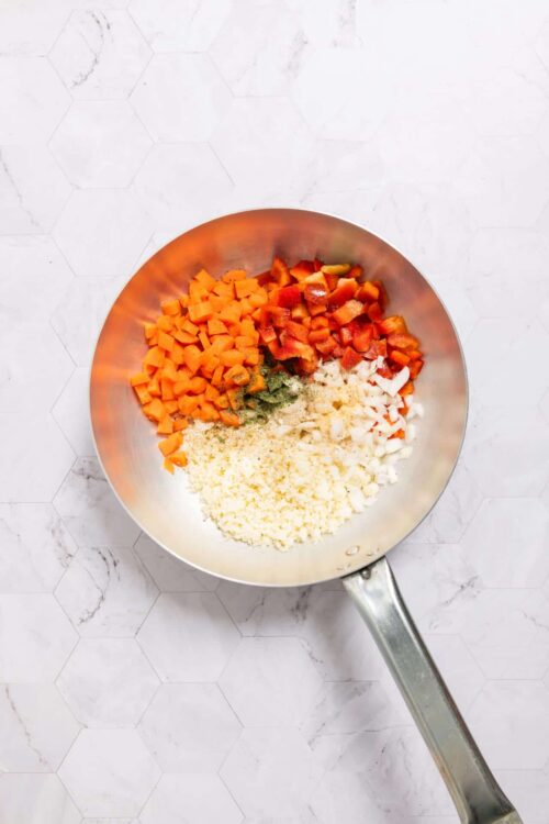 Diced carrots, red bell peppers, onions, and herbs in a stainless steel skillet.