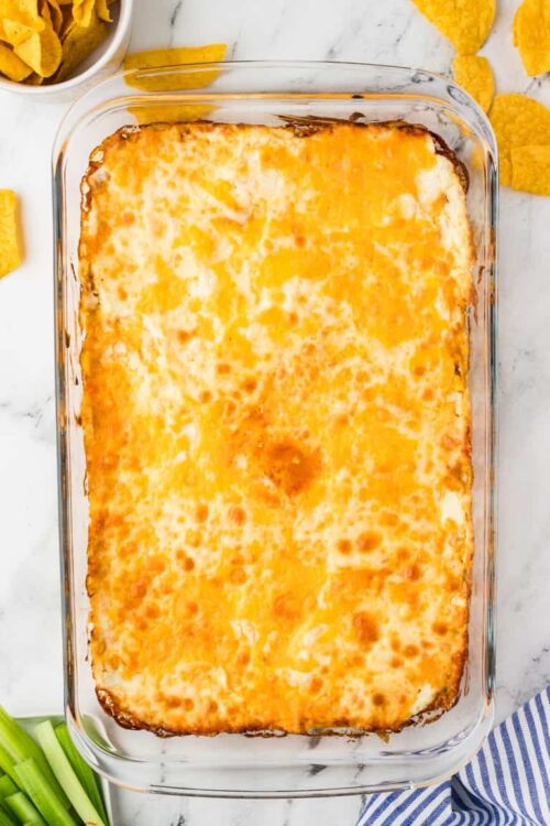 Golden baked cheesy casserole in a glass dish with tortilla chips and celery on a white marble surface.