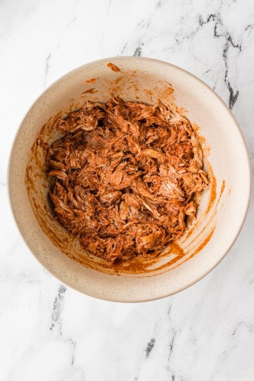 Shredded cooked pork in a creamy tomato sauce in a large beige mixing bowl.