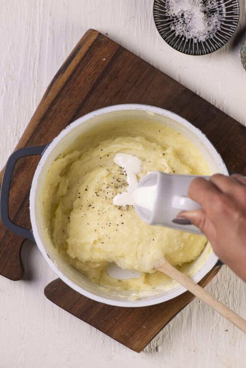 Creamy mashed potatoes being prepared in a pot with black pepper and cream, on a wooden cutting board.