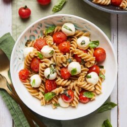 Fresh cherry tomato and mozzarella pasta salad garnished with basil leaves in a white bowl.