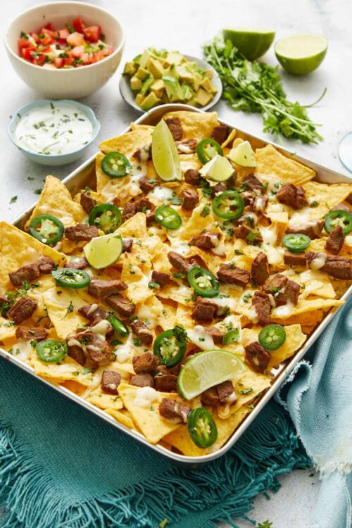 Crispy sheet tray nachos topped with beef, jalapeños, melted cheese, and lime wedges, served with fresh toppings.