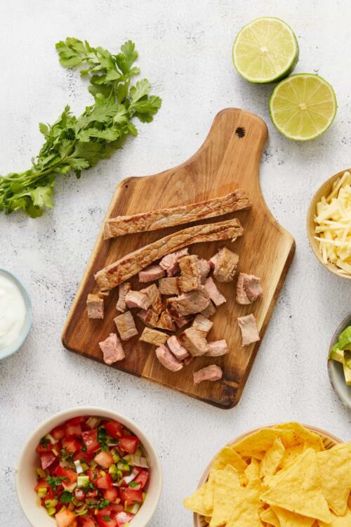 Fresh cilantro leaves on wooden cutting board with cooked pork strips and diced pork, surrounded by chopped tomatoes, tortilla chips, lime halves, and shredded cheese, perfect for Mexican tacos.