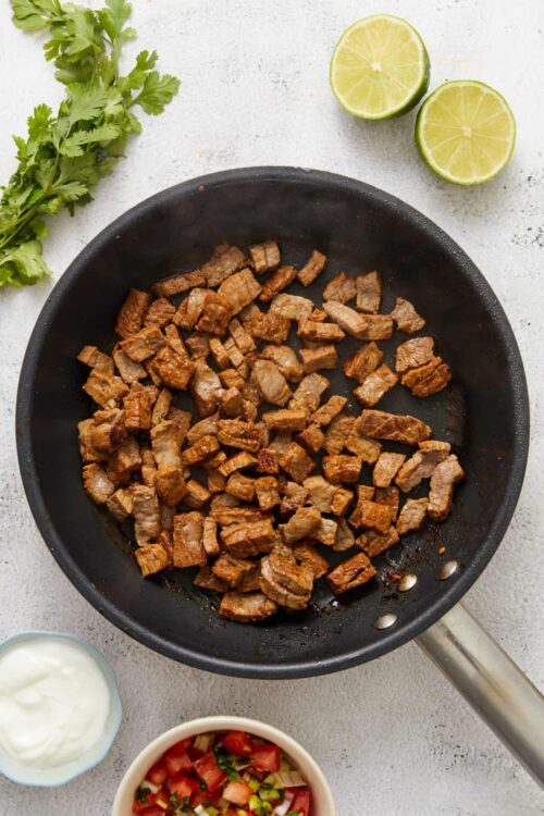 Sautéed beef chunks in a black skillet with lime and fresh herbs on a white background.