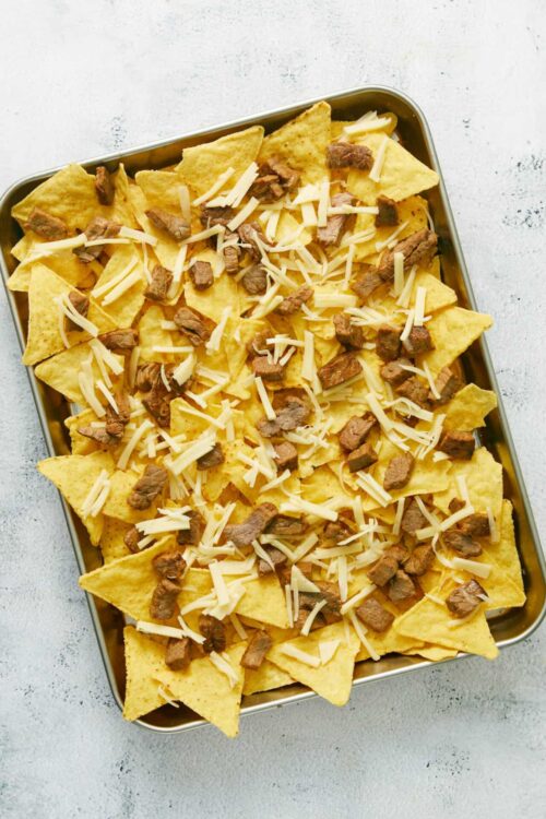 Crispy baked nachos topped with melted cheese and seasoned steak, served in a metal tray.