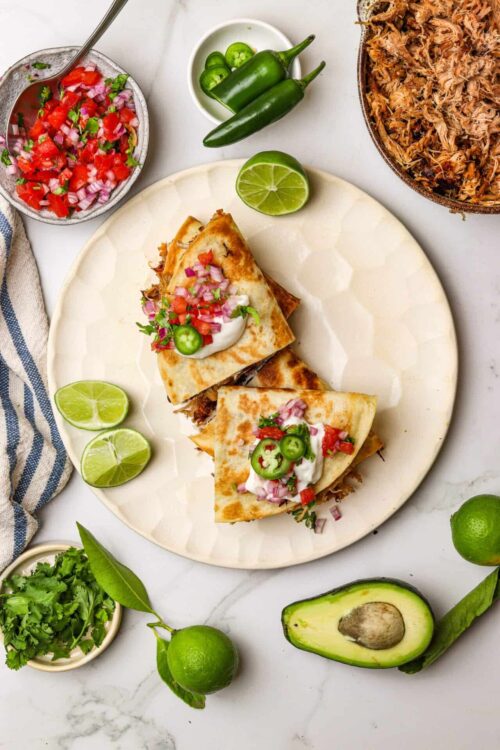 Fresh carnitas tacos with salsa and lime, accompanied by avocado, cilantro, and jalapenos on a white marble surface.