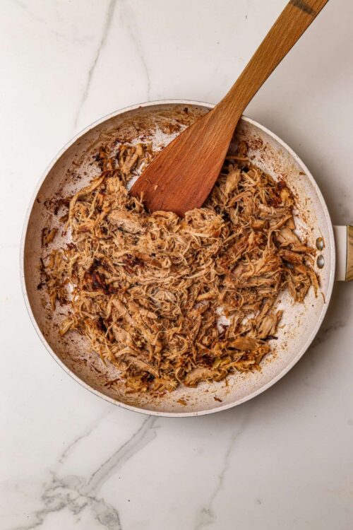 Shredded cooked chicken in a white skillet with a wooden spatula on a white marble surface.