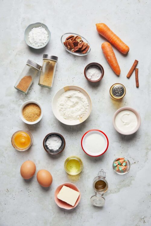 Fresh carrot cake ingredients on a white surface, including carrots, eggs, pecans, spices, and flour.