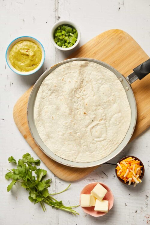 Flour tortilla on a skillet with chopped green onions, shredded cheese, butter, sour cream, and salsa on a white wooden surface.