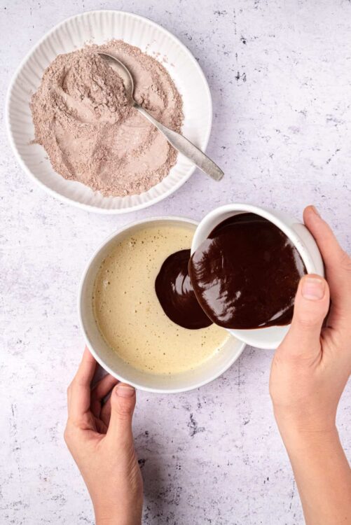 Cream-colored mix being poured into a bowl of chocolate ganache, with a small dish of cocoa powder nearby.