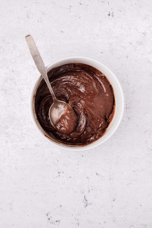 Silky chocolate frosting in a white bowl with a spoon, perfect for baking and dessert decoration.