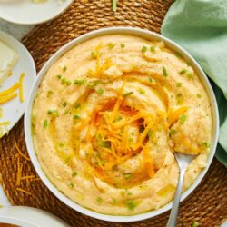Creamy mashed potatoes topped with shredded cheese and chopped chives, served in a white bowl on a woven placemat.