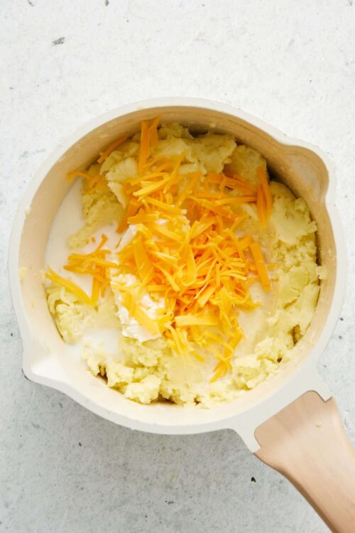 Creamy mashed potatoes with shredded cheddar cheese, prepared in a white mixing bowl.