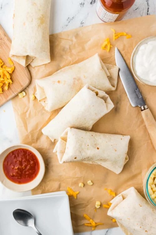 Wrapped cheese and chicken burritos on parchment paper with salsa and sour cream; ready for baking.
