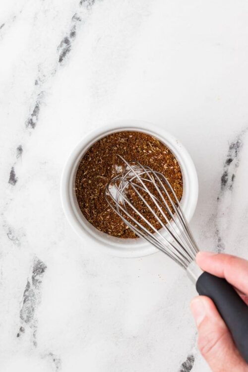Ground spice blend in white bowl, whisk ready for mixing, on a marble surface.
