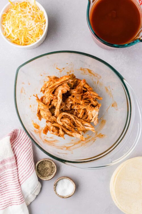Shredded cooked chicken in glass mixing bowl with spices and ingredients around, ready for a recipe.