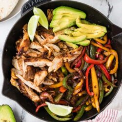 Grilled chicken with sautéed colorful bell peppers, sliced avocado, and lime wedges in a cast iron skillet.