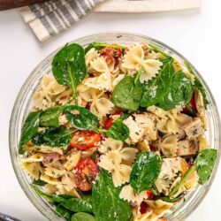 Roasted chicken pasta salad with cherry tomatoes and fresh spinach in a glass bowl.