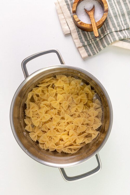Cooked pasta in a metal pot on a white surface with salt and a striped cloth nearby.