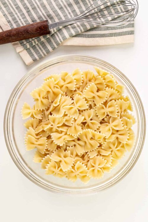 Raw uncooked fusilli pasta in a glass bowl on white background with a whisk and kitchen towel nearby.