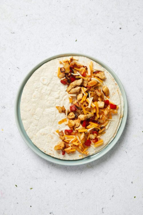 Sliced flour tortillas topped with cooked chicken, red bell peppers, shredded cheese, and sautéed vegetables for chicken fajitas.