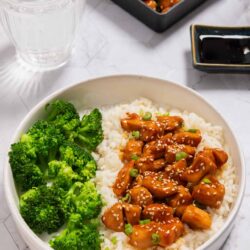 Savory chicken with rice and broccoli in a white bowl, with soy sauce and sweet and sour chicken in the background.