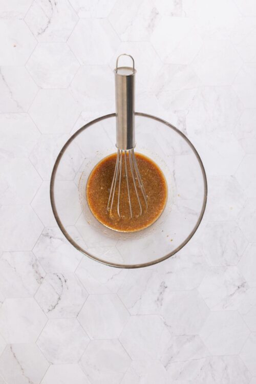 Smooth brown sauce being whisked in a glass bowl with a metal whisk attachment, on a white marble surface.