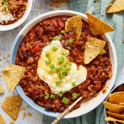 Cheesy beef chili with toppings, sour cream, and tortilla chips in a white bowl for a hearty comfort food meal.