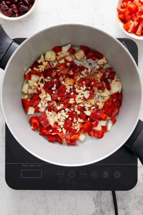 Chopped onions, garlic, and red bell peppers sautéing in a pot on a stovetop.
