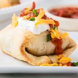 Soft taco with shredded cheese, sour cream, and bacon on side, served on white plate.