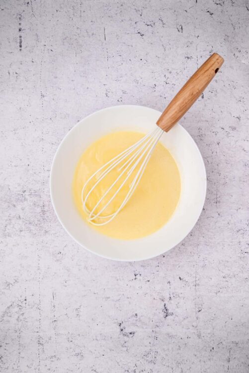 Fluffy yellow cake batter in a white bowl with a whisk and wooden handle, ready to bake.