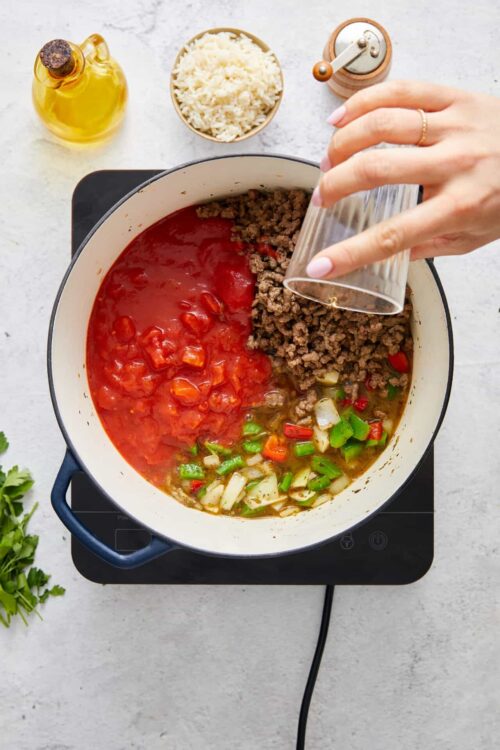 Ground beef being added to a skillet with diced vegetables and tomato sauce for a savory recipe.