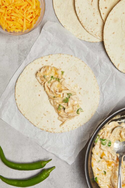 Cheese and chicken quesadilla being assembled on a flour tortilla with shredded cheese nearby.