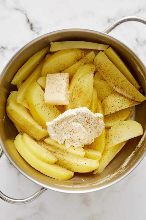 Freshly sliced potatoes with seasoning, butter, and cream in a stainless steel pot for stewing or mash preparation.