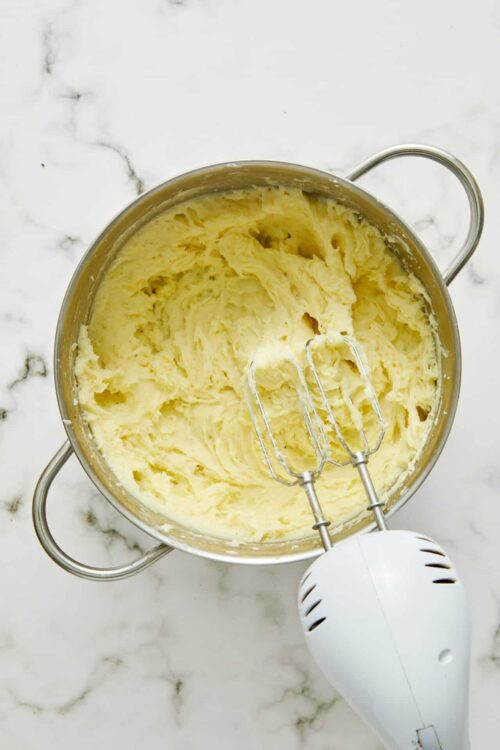 Creamy mashed potatoes in a stainless steel mixing bowl with beaters and a handheld mixer on white marble surface.