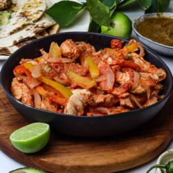 Hearty chicken fajitas with colorful peppers on a black bowl, surrounded by fresh lime, avocado, and traditional accompaniments.