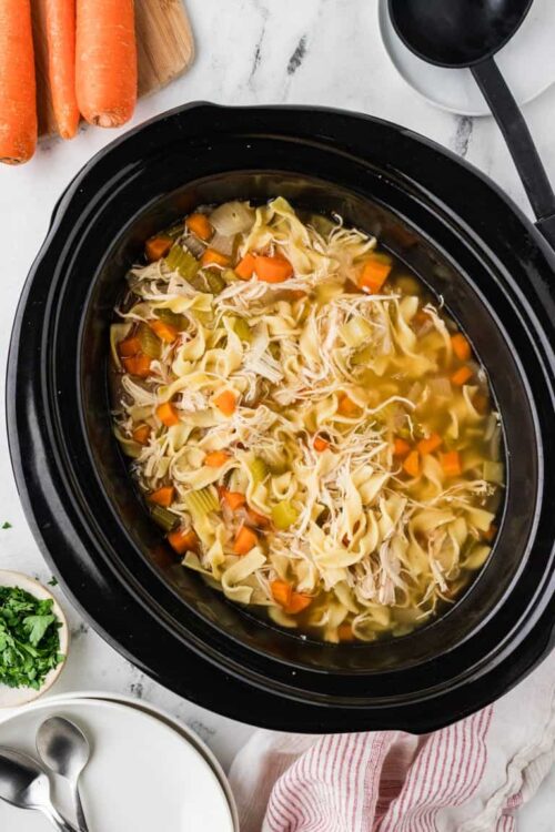 Slow cooker chicken noodle soup with vegetables and pasta in black crockpot.