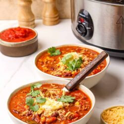 Savory bowls of homemade chili topped with cheese and cilantro, served next to a slow cooker on a kitchen countertop.