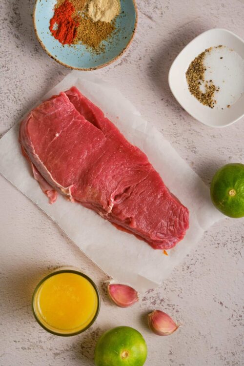 Fresh raw beef tenderloin for recipe; assorted spices, garlic, lime, and seasonings for cooking; Baked Bree food blog ingredients.