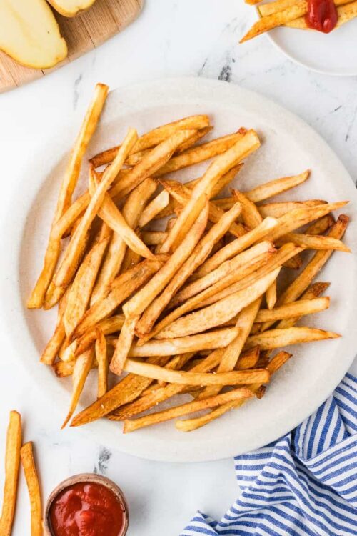 Golden crispy homemade French fries on a white plate, served with ketchup and fresh potatoes on a marble surface.