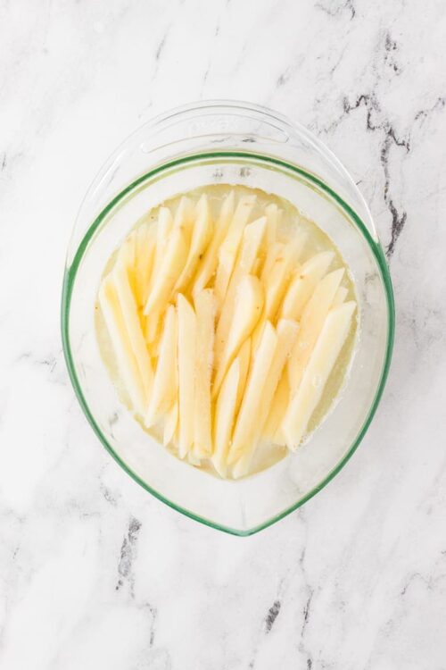 Sliced cheese soaking in water in a glass mixing bowl on a white marble surface.
